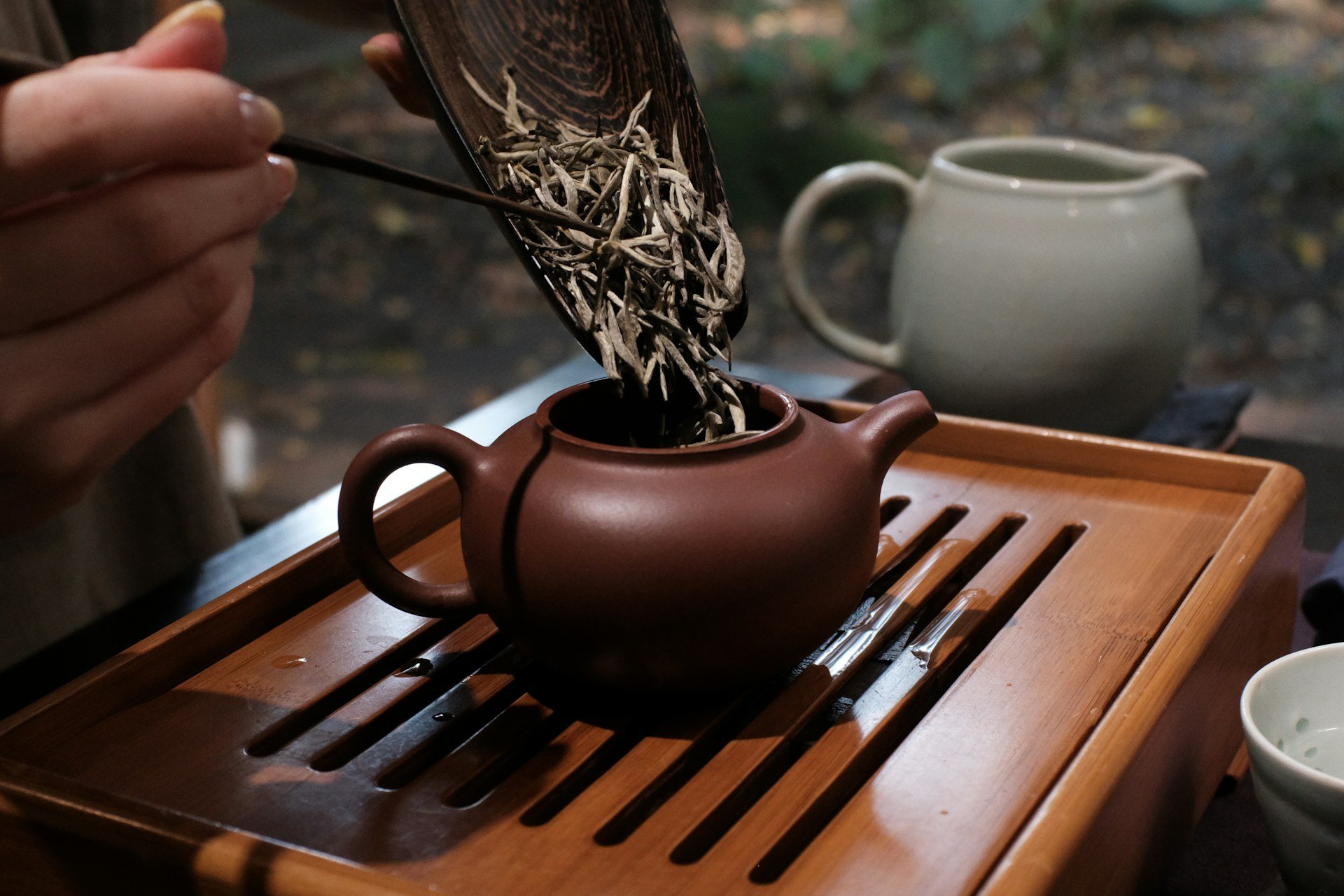 Loose white tea leaves being slid into a clay teapot on a wooden tea tray