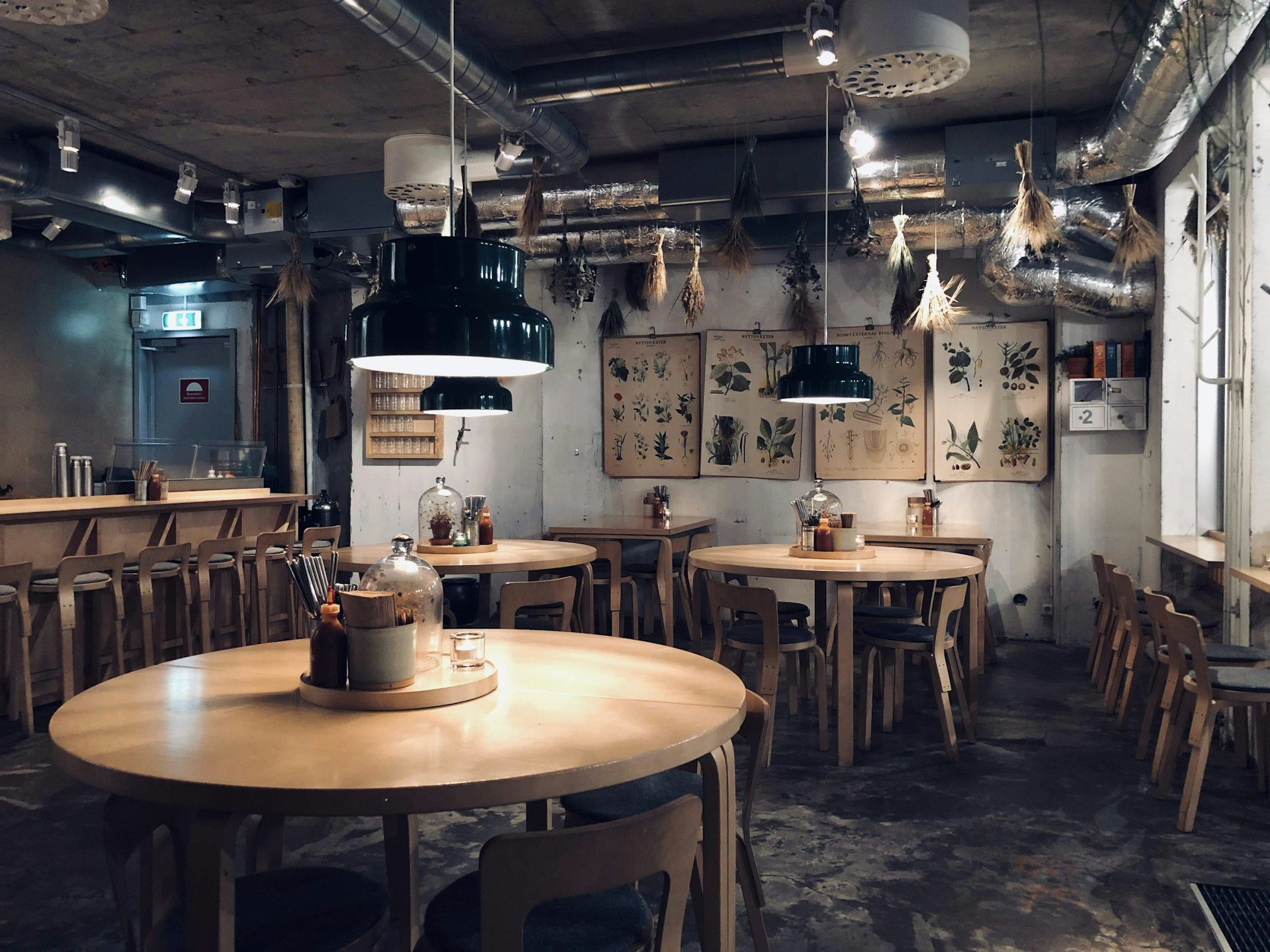 A quiet cafe with round wooden tables, bentwood chairs and pendant lamps under botanical prints